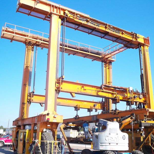 Operator under a yellow mobile gantry crane lifting fabricated structure outdoors at customer site