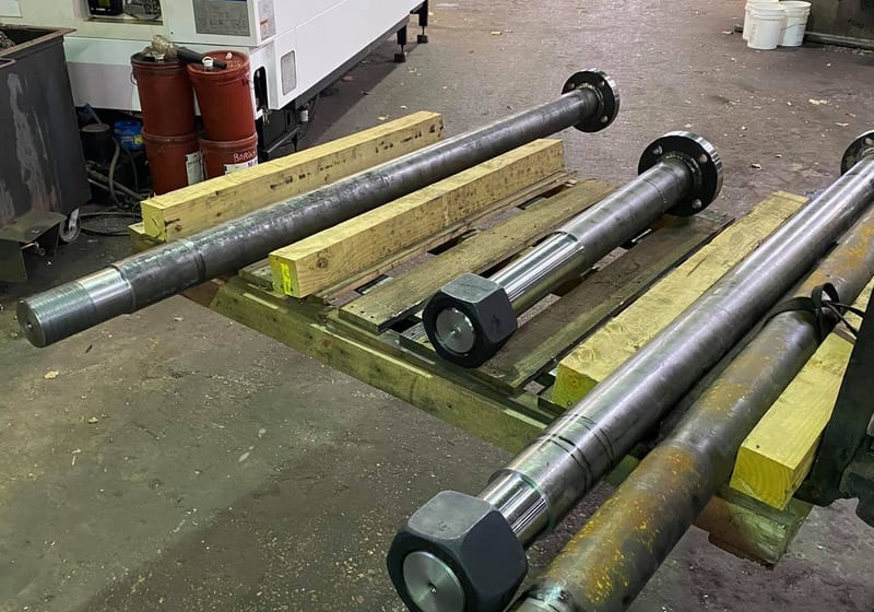 Four large custom-machined steel shafts on a pallet in front of a Mazak CNC lathe at A&K Machine & Fabrication