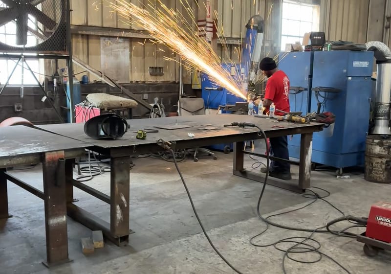 Welder/grinder making sparks on structural beam, blue dust collector + Lincoln welder behind, full shop floor context — A&K Machine & Fabrication, Jacksonville FL