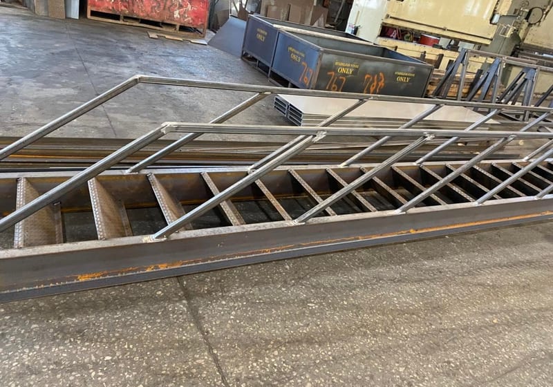 Large fabricated steel ship's ladder with angled stringers and safety railing on shop floor at A&K