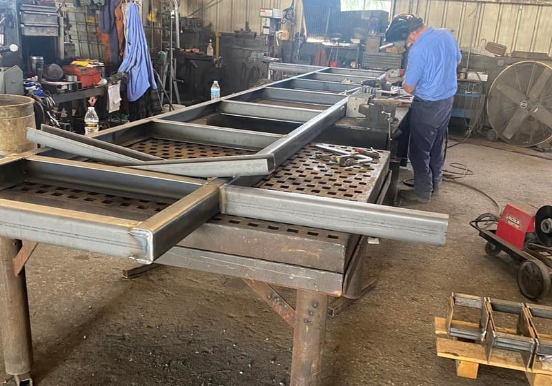 Fabricator welding a large steel trailer frame on welding table with American flag at A&K